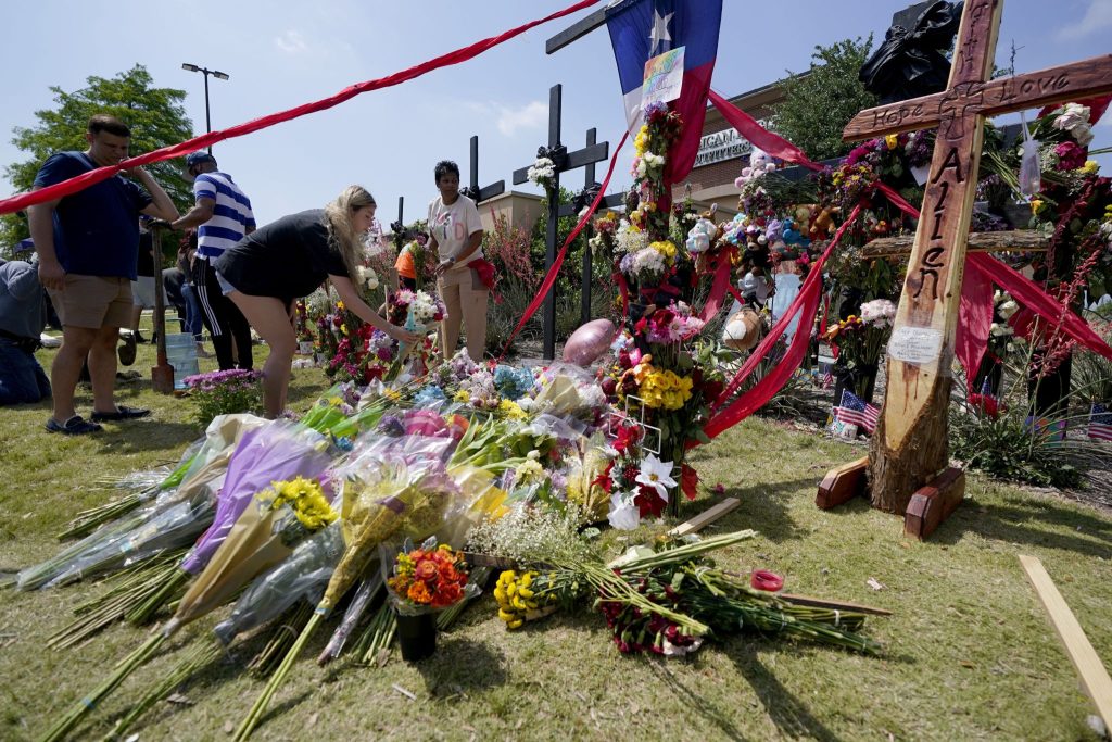 Visitors to a makeshift memorial at the Allen Premium Outlets in Texas leave flowers in front of a large cross. The cross has the words "Hope, Love, Allen" engraved into it.