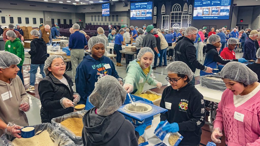 Volunteers chant “PROTEIN! VEGETABLES! SOY! RICE!” as they scoop ingredients into a MannaPack bag in the auditorium of The Park Church of Christ.