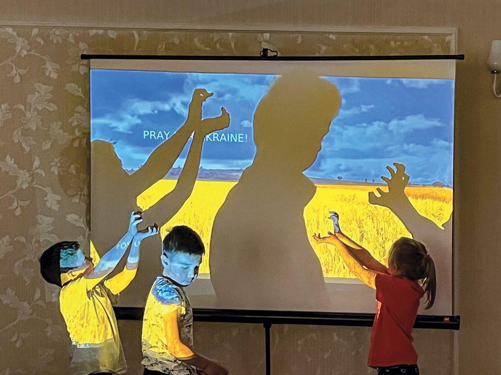 As they get ready to watch a movie in the hotel, Ukrainian children make shadow puppets on a projected computer screen that reads “Pray for Ukraine!”